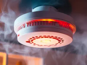 Runder Rauchmelder an der Decke mit rot leuchtendem Alarmlicht, umgeben von aufsteigendem Rauch.