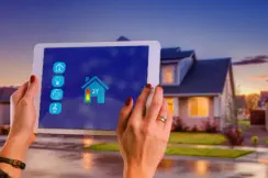 Hände halten ein Tablet vor einem modernen Einfamilienhaus. Auf dem Bildschirm ist eine Smart-Home-Anwendung mit einem Haus-Symbol und verschiedenen Steuerungsoptionen zu sehen.